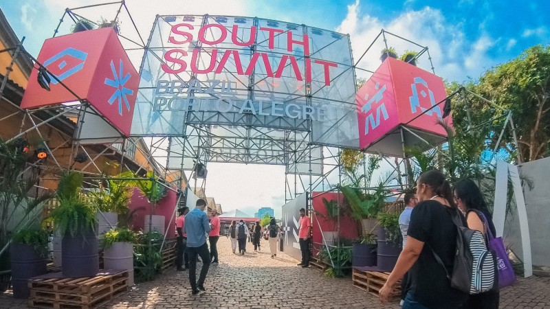 A foto mostra cerca de dez pessoas cruzando o portal da entrada do evento, no qual consta, em rosa, "South Summit Brazil Porto Alegre".