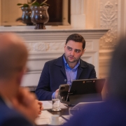 Há três pessoas na foto sentadas à mesa. Duas estão de costas e desfocadas. Gabriel Souza aparece focado de frente ao fundo, no centro da imagem.