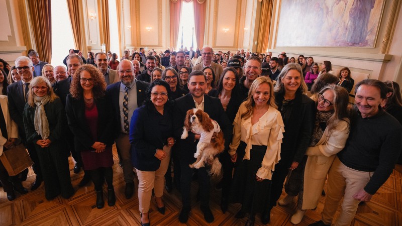 A foto mostra um grupo grande e diverso de pessoas, incluindo homens e mulheres, posando para uma foto em um sal&atilde;o espa&ccedil;oso com um teto alto e um grande afresco em uma das paredes. No centro, o governador em exerc&iacute;cio, Gabriel Souza, est&aacute; segurando um c&atilde;o de porte m&eacute;dio, de pelagem branca e caramelo, que veste uma camiseta branca. Ao lado de Gabriel, h&aacute; um grupo de pessoas sorridentes, incluindo a Secret&aacute;ria do Meio Ambiente e Infraestrutura, Marjorie Kauffmann, de blusa branca. A plateia, formada por muitas pessoas, est&aacute; em p&eacute; no fundo. O piso &eacute; de madeira e a ilumina&ccedil;&atilde;o &eacute; natural.