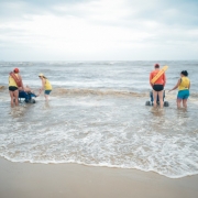 Foto em ambiente aberto do mar, com duas pessoas em cadeiras híbridas entrando na água, monitoradas por dois guarda-vidas. Elas aparecem de costas.

