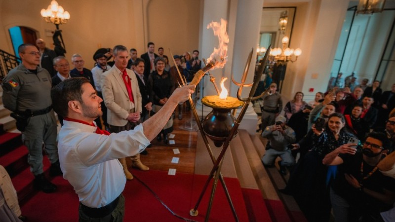 Gabriel Souza aparece à esquerda da imagem acendendo o Candeeiro Crioulo, que está ao centro da foto. Ao redor, cerca de 20 pessoas observam a cena.