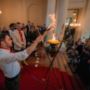 Gabriel Souza aparece à esquerda da imagem acendendo o Candeeiro Crioulo, que está ao centro da foto. Ao redor, cerca de 20 pessoas observam a cena.