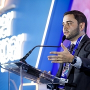 Homem em traje formal fala ao microfone no palco da conferência, com tela iluminada ao fundo exibindo nome do evento. Ambiente profissional e moderno.