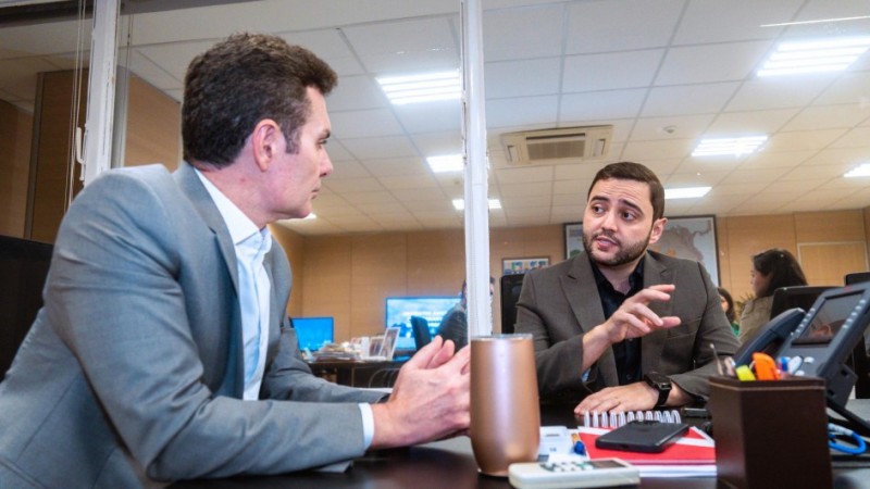 Dois homens sentados em uma mesa de escritório conversam, em ambiente iluminado por luzes de teto. O homem à esquerda veste terno cinza e camisa clara, enquanto o da direita usa terno escuro e gesticula com a mão durante a fala. Sobre a mesa, há objetos de escritório, como canetas, caderno, telefone e uma garrafa térmica. Ao fundo, outras pessoas trabalham em suas mesas.
