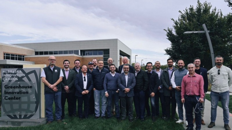 Uma foto externa de um grupo de cerca de 20 homens de pé, posando na frente de um letreiro e um prédio. O letreiro de cor escura, no canto inferior esquerdo, diz "NEBRASKA INNOVATION CAMPUS" e, abaixo, "Greenhouse Innovation Center". O prédio atrás é moderno, de dois andares, com fachada em cores claras e muitas janelas. Os homens estão vestidos de maneira casual de negócios ou casual e parecem ser membros de uma comitiva, possivelmente na área de agricultura ou tecnologia de estufas, em visita ao centro de pesquisa no Nebraska, EUA. O céu está nublado.