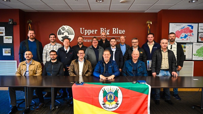 Grupo de 17 homens posa para foto em uma sala de reuniões com parede avermelhada, onde se lê “Upper Big Blue Natural Resources District”. À frente da mesa, uma bandeira do Rio Grande do Sul está estendida. O vice-governador Gabriel Souza aparece ao centro, sentado, com jaqueta azul escura.