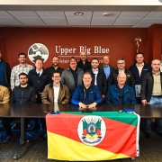 Grupo de 17 homens posa para foto em uma sala de reuniões com parede avermelhada, onde se lê “Upper Big Blue Natural Resources District”. À frente da mesa, uma bandeira do Rio Grande do Sul está estendida. O vice-governador Gabriel Souza aparece ao centro, sentado, com jaqueta azul escura.