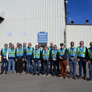 A imagem apresenta um grupo de visitantes posicionado em frente a um grande galpão industrial de paredes metálicas claras. Todos vestem coletes de segurança fluorescentes com a inscrição “VISITOR”.