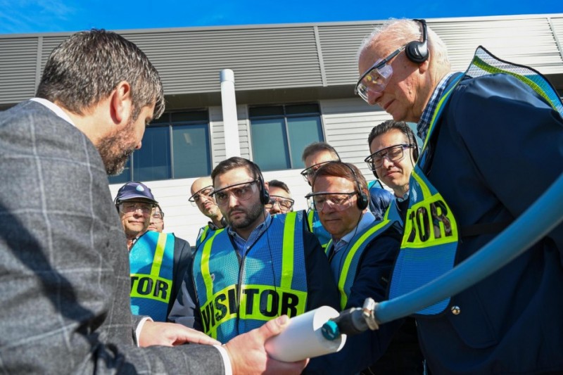 A foto mostra um grupo de dez pessoas em um ambiente externo. Todas, exceto uma, usam coletes de segurança fluorescentes com a palavra “VISITOR” e óculos de proteção. Ao centro, encontra-se o vice-governador Gabriel Souza. À esquerda, um homem grisalho que veste um terno xadrez segura uma mangueira.