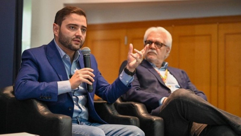 Dois homens sentados em poltronas pretas participam de conversa formal. Um fala ao microfone com expressão séria, o outro observa atento. Ambos usam blazer azul e jeans. Ambiente interno e profissional.