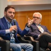 Dois homens sentados em poltronas pretas participam de conversa formal. Um fala ao microfone com expressão séria, o outro observa atento. Ambos usam blazer azul e jeans. Ambiente interno e profissional.