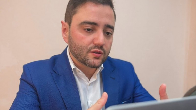 Uma foto em plano médio de um homem de pele clara, cabelo escuro e barba curta, vestido com um blazer azul vibrante sobre uma camisa social branca. Ele está sentado em uma mesa e gesticula com as mãos enquanto olha para frente, possivelmente para uma tela de notebook parcialmente visível no canto inferior direito. O fundo é de cor bege claro, liso e discreto. A iluminação é suave, destacando o rosto e o blazer do homem.