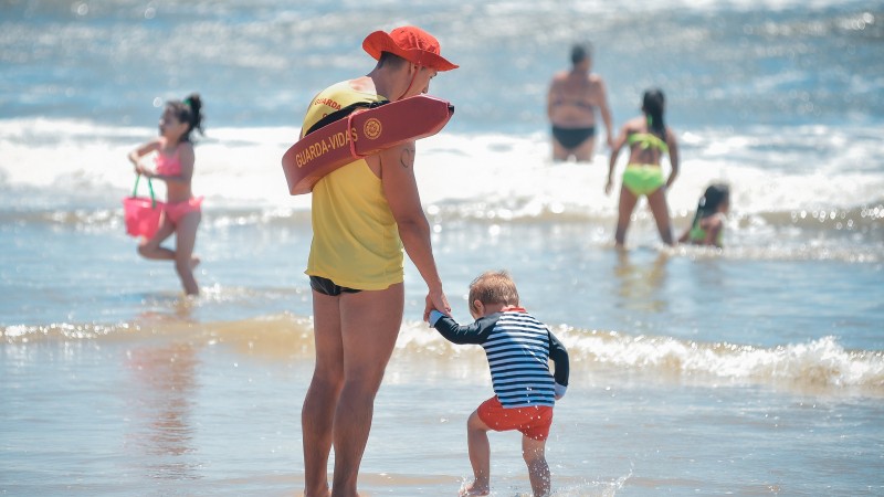 A imagem mostra um guarda-vida de costas, segurando a mão de uma criança. Ele está de frente para o mar, onde três crianças e um adulto se banham.