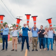 Uma foto externa de um grupo de cerca de 18 homens posando para a câmera em frente a um prédio moderno, possivelmente em um campus de pesquisa ou inovação. A maioria dos homens veste roupas casuais de negócios, como blazers, camisas polo ou casacos leves. O grupo está reunido em frente a uma placa de sinalização de metal cinza com o logotipo e o nome: "NEBRASKA INNOVATION CAMPUS" e "Greenhouse Innovation Center" em letras grandes, indicando o local da visita. O edifício ao fundo é moderno, com fachada de cor clara (cinza claro e bege) e amplas janelas horizontais escuras. O gramado em primeiro plano está verde e há uma árvore frondosa e uma luminária de rua no canto direito. O céu está nublado, e a comitiva parece estar em uma missão de pesquisa em irrigação ou tecnologia agrícola nos EUA.
