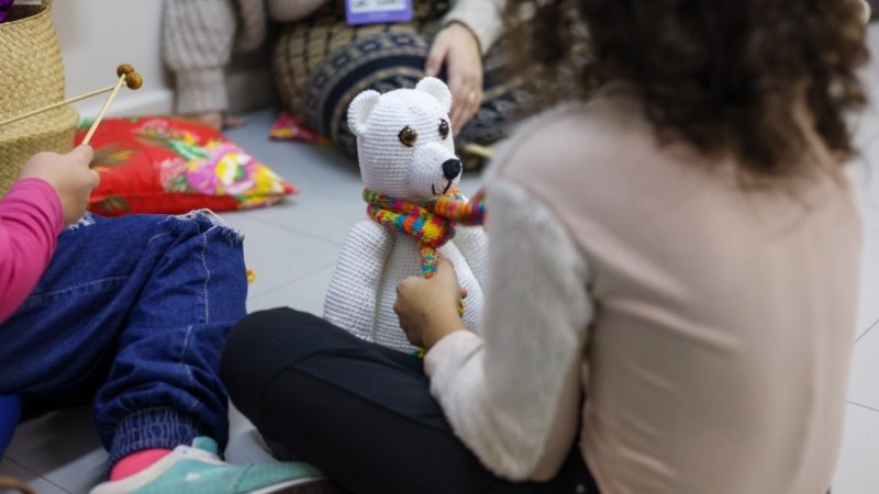 A imagem mostra uma crian&ccedil;a de costas, com o cabelo cacheado. Ela est&aacute; sentada no ch&atilde;o, brincando com um urso branco de cachecol colorido. Seu rosto n&atilde;o est&aacute; vis&iacute;vel. Ao lado, aparecem as pernas de outra crian&ccedil;a sentada perto da menina e de uma pessoa adulta com crach&aacute;.