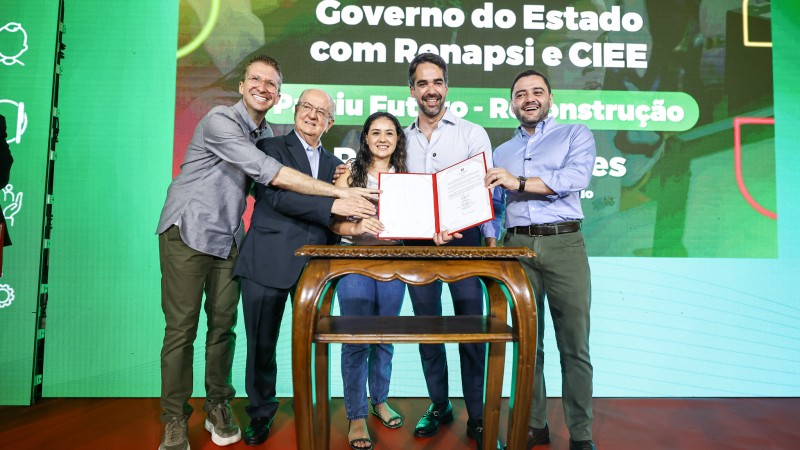Uma fotografia em um evento interno, com fundo verde-claro e um letreiro grande na parte superior. Quatro homens e uma mulher est&atilde;o ao redor de uma pequena mesa de madeira escura com tampo oval, no centro da imagem. Eles est&atilde;o em p&eacute;, sorrindo e estendendo as m&atilde;os para um documento que est&aacute; sendo segurado pela mulher. A mulher, vestindo uma cal&ccedil;a jeans escura e uma blusa azul-claro, est&aacute; no centro da mesa, segurando o documento vermelho (uma pasta ou certificado). O homem &agrave; sua direita (esquerda na foto), mais velho, de terno cinza, apoia a m&atilde;o sobre a dela. O homem &agrave; esquerda (direita na foto), de camisa clara com as mangas arrega&ccedil;adas, tamb&eacute;m apoia a m&atilde;o sobre a da mulher. Outros dois homens, um de camisa cinza (na extrema esquerda) e outro de camisa azul-escura (na extrema direita), est&atilde;o olhando para a c&acirc;mera e sorrindo, com as m&atilde;os espalmadas ou levemente levantadas, pr&oacute;ximos ao grupo central. O letreiro acima diz: "Assinatura de Parceria - Governo do Estado com Renapsi e CIEE" e logo abaixo, em letras maiores: "Partiu Futuro - Reconstru&ccedil;&atilde;o". A ilumina&ccedil;&atilde;o &eacute; clara e focada no grupo de pessoas.