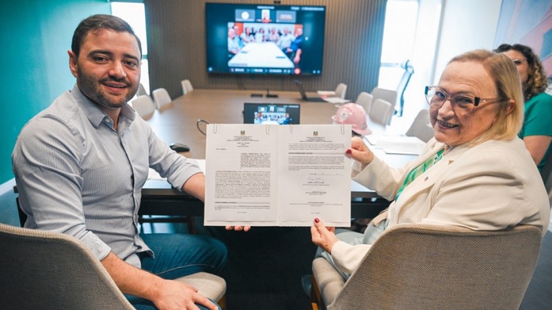 Vice governador Gabriel Souza aparece na foto ao lado da secret&aacute;ria Arita Bergmann, ambos seguram um documento, com uma tela com outros participantes online ao fundo.