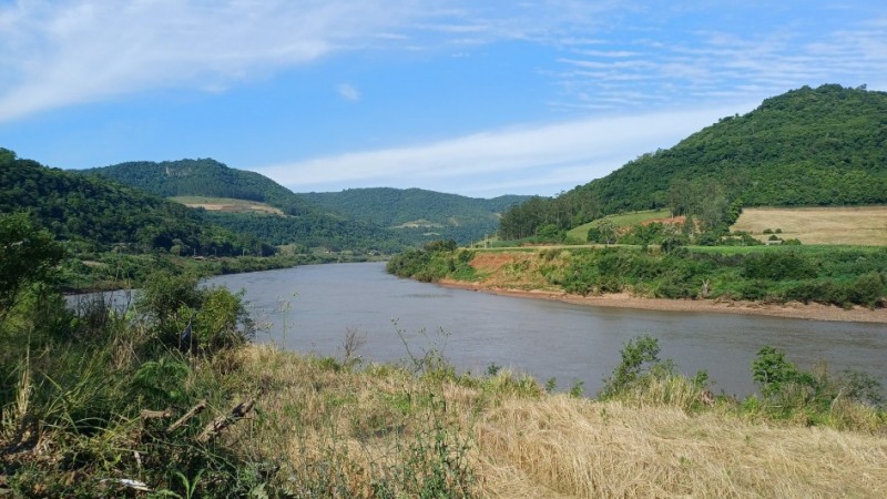 A fotografia apresenta uma vista panor&acirc;mica de um rio largo que serpenteia por um vale entre colinas cobertas de vegeta&ccedil;&atilde;o, sob um c&eacute;u azul claro. Detalhes da Paisagem O Rio: O curso d'&aacute;gua ocupa a parte central da imagem, apresentando &aacute;guas de tom acastanhado e uma superf&iacute;cie relativamente calma que reflete a claridade do dia. O Relevo: Ao fundo e nas laterais, elevam-se morros e colinas cobertos por uma mata verde e densa. Algumas &aacute;reas nas encostas mostram clar&otilde;es de terra e campos cultivados, evidenciando o uso agr&iacute;cola da terra na regi&atilde;o. Primeiro Plano: Na parte inferior da foto, v&ecirc;-se a vegeta&ccedil;&atilde;o da margem, composta por arbustos verdes, gram&iacute;neas secas de cor amarelada e um solo pedregoso no canto esquerdo. O C&eacute;u: O c&eacute;u est&aacute; bem aberto e azul, com nuvens brancas finas e alongadas que se espalham de forma suave. Contexto De acordo com o t&iacute;tulo do arquivo, a imagem ilustra a implementa&ccedil;&atilde;o de um novo servi&ccedil;o de monitoramento pelo Governo do Estado do Rio Grande do Sul, projetado para prever o n&iacute;vel dos rios e auxiliar na preven&ccedil;&atilde;o de enchentes.