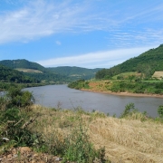A fotografia apresenta uma vista panor&acirc;mica de um rio largo que serpenteia por um vale entre colinas cobertas de vegeta&ccedil;&atilde;o, sob um c&eacute;u azul claro. Detalhes da Paisagem O Rio: O curso d'&aacute;gua ocupa a parte central da imagem, apresentando &aacute;guas de tom acastanhado e uma superf&iacute;cie relativamente calma que reflete a claridade do dia. O Relevo: Ao fundo e nas laterais, elevam-se morros e colinas cobertos por uma mata verde e densa. Algumas &aacute;reas nas encostas mostram clar&otilde;es de terra e campos cultivados, evidenciando o uso agr&iacute;cola da terra na regi&atilde;o. Primeiro Plano: Na parte inferior da foto, v&ecirc;-se a vegeta&ccedil;&atilde;o da margem, composta por arbustos verdes, gram&iacute;neas secas de cor amarelada e um solo pedregoso no canto esquerdo. O C&eacute;u: O c&eacute;u est&aacute; bem aberto e azul, com nuvens brancas finas e alongadas que se espalham de forma suave. Contexto De acordo com o t&iacute;tulo do arquivo, a imagem ilustra a implementa&ccedil;&atilde;o de um novo servi&ccedil;o de monitoramento pelo Governo do Estado do Rio Grande do Sul, projetado para prever o n&iacute;vel dos rios e auxiliar na preven&ccedil;&atilde;o de enchentes.