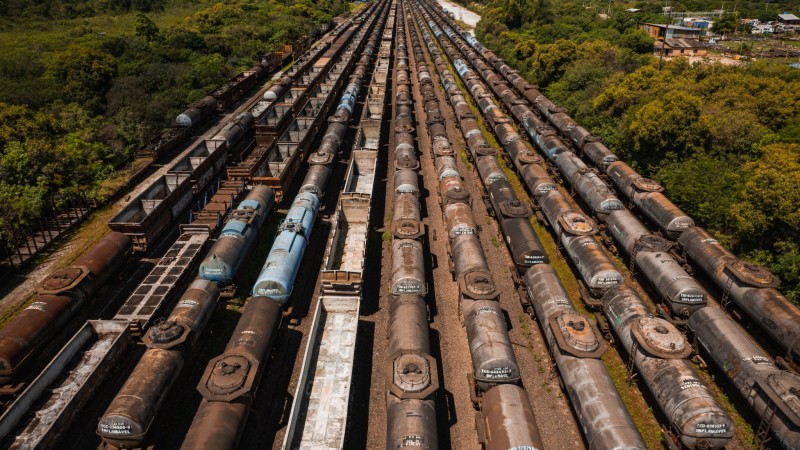 Foto horizontal a&eacute;rea mostra onze fileiras de vag&otilde;es de trem envelhecidos parados lado a lado, em longas fileiras paralelas que se estendem at&eacute; o horizonte. As vias ferrovi&aacute;rias est&atilde;o cercadas por vegeta&ccedil;&atilde;o verde densa &agrave; direita e &agrave; esquerda da imagem. Entre a vegeta&ccedil;&atilde;o que aparece ao fundo, h&aacute; constru&ccedil;&otilde;es baixas. O c&eacute;u est&aacute; claro e o sol ilumina a cena.