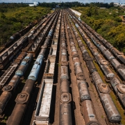 Foto horizontal a&eacute;rea mostra onze fileiras de vag&otilde;es de trem envelhecidos parados lado a lado, em longas fileiras paralelas que se estendem at&eacute; o horizonte. As vias ferrovi&aacute;rias est&atilde;o cercadas por vegeta&ccedil;&atilde;o verde densa &agrave; direita e &agrave; esquerda da imagem. Entre a vegeta&ccedil;&atilde;o que aparece ao fundo, h&aacute; constru&ccedil;&otilde;es baixas. O c&eacute;u est&aacute; claro e o sol ilumina a cena.