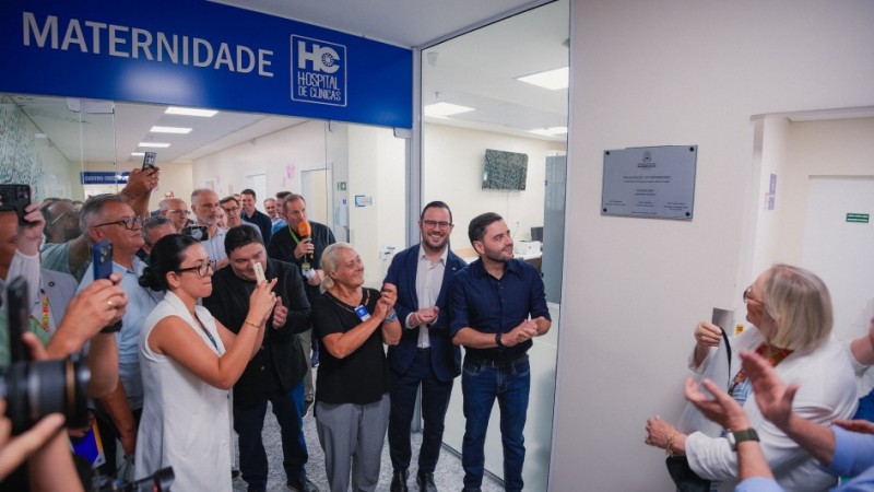 Foto horizontal mostra um grupo de cerca de 20 pessoas reunidas em um corredor iluminado de um hospital, diante da entrada de uma &aacute;rea identificada por uma placa azul no alto com o texto &ldquo;MATERNIDADE &ndash; HC Hospital de Cl&iacute;nicas&rdquo;. Entre elas, v&ecirc;-se o vice-governador Gabriel Souza e a secret&aacute;ria estadual da Sa&uacute;de, Arita Bergmann, &Agrave; direita da imagem, h&aacute; uma placa inaugural prateada fixada &agrave; parede. As pessoas est&atilde;o posicionadas em semic&iacute;rculo, olhando para a placa. Algumas est&atilde;o com c&acirc;meras ou celulares levantados, outras batendo palmas. V&aacute;rias usam crach&aacute;s ou adesivos no peito. O local tem piso claro e paredes brancas, com portas de vidro e salas iluminadas ao fundo.