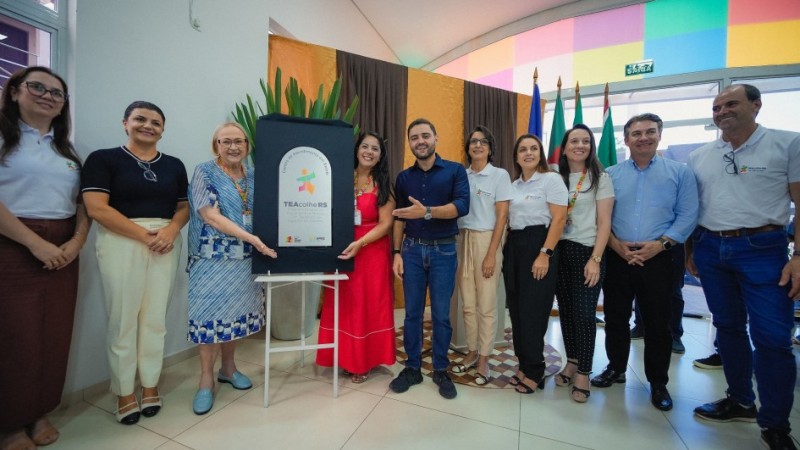 A imagem mostra um grupo de dez pessoas reunidas em um ambiente interno bem iluminado. Entre elas, est&atilde;o o vice-governador Gabriel Souza e a secret&aacute;ria estadual da Sa&uacute;de, Arita Bergmann. As pessoas est&atilde;o posicionadas em semic&iacute;rculo ao redor de um painel vertical apoiado sobre um trip&eacute; branco. Esse painel &eacute; o ponto central da cena e exibe um logotipo colorido com formas geom&eacute;tricas, acompanhado do texto &ldquo;TEAcolhe RS&rdquo;. O espa&ccedil;o onde o grupo est&aacute; apresenta piso claro e paredes com elementos decorativos: atr&aacute;s do painel h&aacute; uma cortina escura e, ao lado, uma parede revestida com cores vibrantes em tons de rosa, azul, verde, amarelo e laranja. &Agrave; direita da imagem, atr&aacute;s das pessoas, est&atilde;o posicionadas v&aacute;rias bandeiras hasteadas. H&aacute; tamb&eacute;m um arranjo de plantas ao fundo, atr&aacute;s do trip&eacute;.