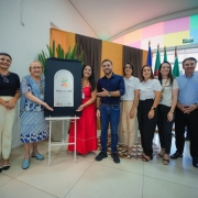 A imagem mostra um grupo de dez pessoas reunidas em um ambiente interno bem iluminado. Entre elas, est&atilde;o o vice-governador Gabriel Souza e a secret&aacute;ria estadual da Sa&uacute;de, Arita Bergmann. As pessoas est&atilde;o posicionadas em semic&iacute;rculo ao redor de um painel vertical apoiado sobre um trip&eacute; branco. Esse painel &eacute; o ponto central da cena e exibe um logotipo colorido com formas geom&eacute;tricas, acompanhado do texto &ldquo;TEAcolhe RS&rdquo;. O espa&ccedil;o onde o grupo est&aacute; apresenta piso claro e paredes com elementos decorativos: atr&aacute;s do painel h&aacute; uma cortina escura e, ao lado, uma parede revestida com cores vibrantes em tons de rosa, azul, verde, amarelo e laranja. &Agrave; direita da imagem, atr&aacute;s das pessoas, est&atilde;o posicionadas v&aacute;rias bandeiras hasteadas. H&aacute; tamb&eacute;m um arranjo de plantas ao fundo, atr&aacute;s do trip&eacute;.