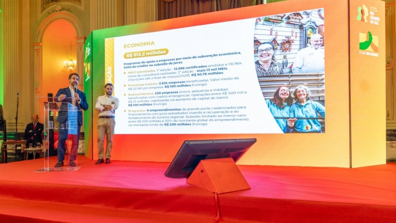 A foto registra um palco amplo, onde o governador e o vice-governador est&atilde;o posicionados &agrave; frente de um tel&atilde;o de grandes dimens&otilde;es. O painel exibe conte&uacute;do sobre Economia, com dados, valores e imagens ilustrativas.