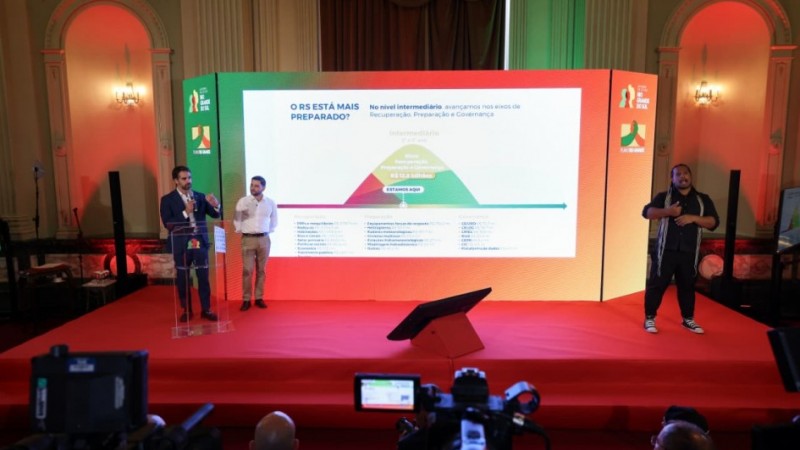 A foto mostra o governador Eduardo Leite e o vice-governador Gabriel Souza em um palco, com um tel&atilde;o ao fundo com informa&ccedil;&otilde;es sobre o Plano Rio Grande.
