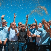 Grupo de cerca de dez pessoas joga gr&atilde;os de soja para o alto em comemora&ccedil;&atilde;o, sob c&eacute;u azul intenso. V&aacute;rios usam chap&eacute;us de palha e camisas claras. No centro, destaca-se um homem de preto. A cena celebra a abertura da colheita da soja.