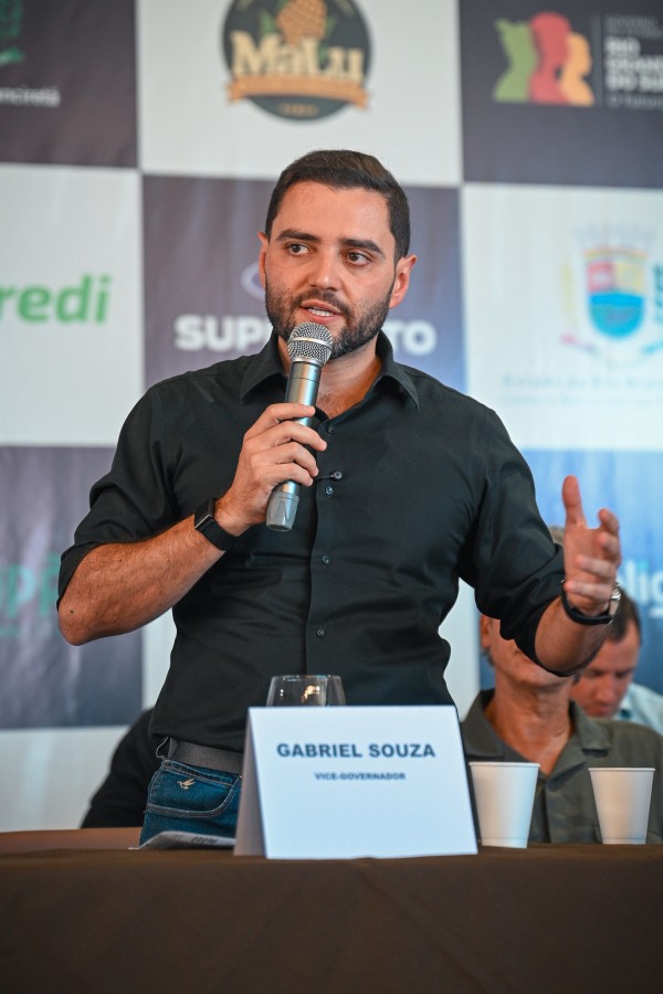 Homem jovem de camisa preta discursa ao microfone com expressão animada e gesto com a mão. À sua frente, plaquinha com "Gabriel Souza – Vice-Governador". Ao fundo, logos de empresas.