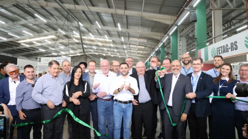 A foto registra uma cerim&ocirc;nia de inaugura&ccedil;&atilde;o em pavilh&atilde;o coberto. Um grupo grande est&aacute; alinhado segurando uma fita verde para o corte simb&oacute;lico, com o vice-governador ao centro. O local possui estandes ao fundo e ilumina&ccedil;&atilde;o t&iacute;pica de galp&atilde;o de eventos, com estrutura met&aacute;lica e corredores amplos.