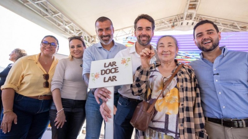 Foto em ambiente externo, sob uma estrutura coberta. Sete pessoas adultas est&atilde;o em p&eacute;, lado a lado, de frente para a c&acirc;mera. No centro, o governador Eduardo Leite segura uma placa branca com o texto &ldquo;LAR DOCE LAR&rdquo; e desenhos de flores, enquanto uma senhora, ao seu lado, exibe uma chave erguida. Ao fundo, h&aacute; um painel colorido em tons de roxo e azul e a estrutura do evento.