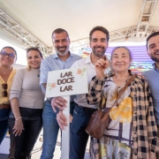 Foto em ambiente externo, sob uma estrutura coberta. Sete pessoas adultas est&atilde;o em p&eacute;, lado a lado, de frente para a c&acirc;mera. No centro, o governador Eduardo Leite segura uma placa branca com o texto &ldquo;LAR DOCE LAR&rdquo; e desenhos de flores, enquanto uma senhora, ao seu lado, exibe uma chave erguida. Ao fundo, h&aacute; um painel colorido em tons de roxo e azul e a estrutura do evento.