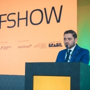 Vice-Governador Gabriel Souza fala ao microfone em um p&uacute;lpito durante um evento, vestindo terno e crach&aacute;. Ao fundo, um painel laranja exibe &ldquo;BFSHOW&rdquo; e logotipos institucionais, com ilumina&ccedil;&atilde;o profissional e luzes verdes laterais.
